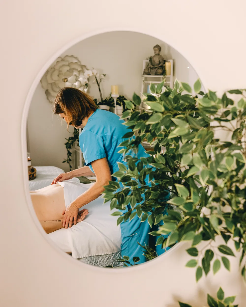 A round mirror reflecting a Revive massage treatment, framed by green plants.
