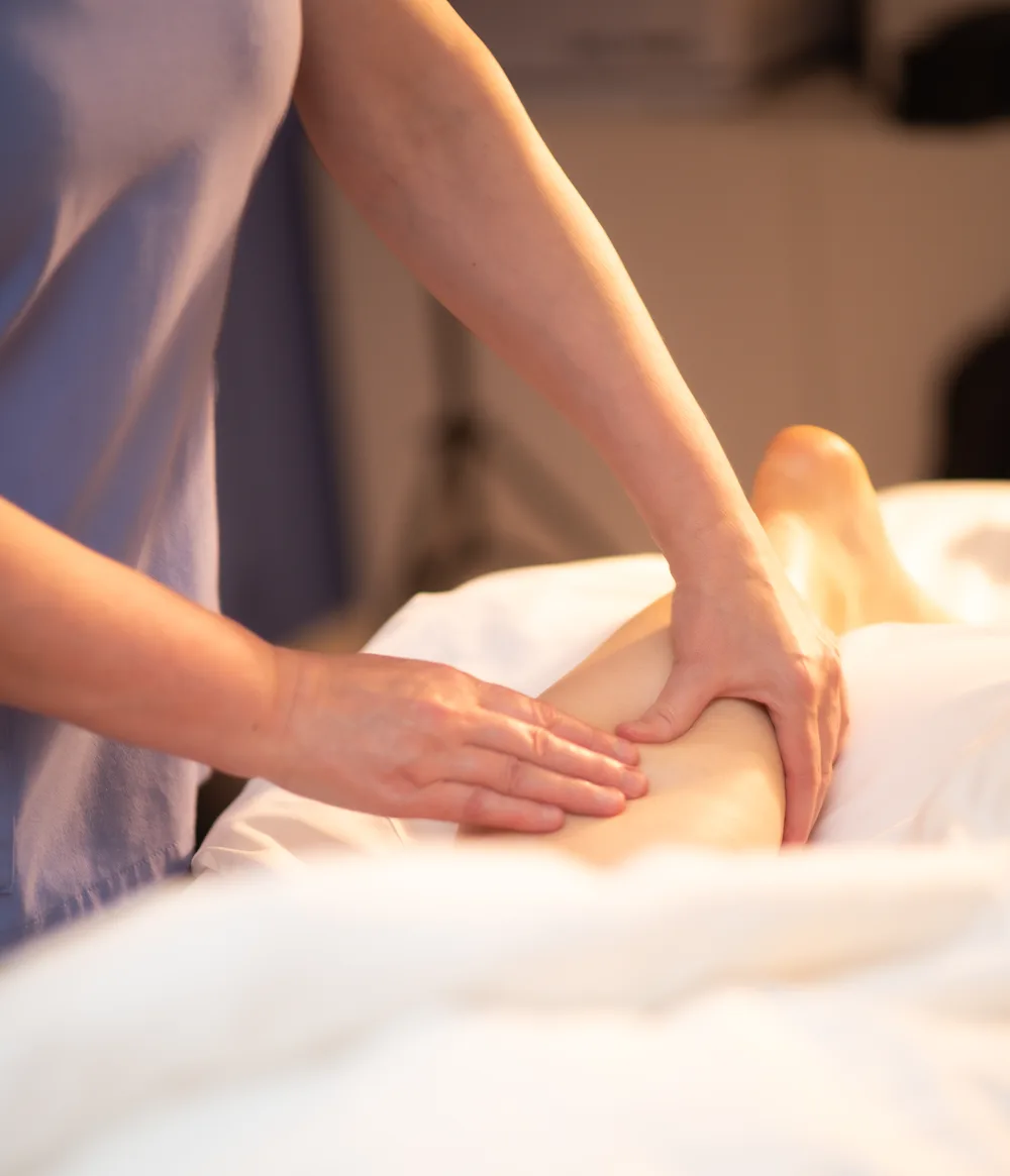 A therapist using her hands on a client’s lower leg on a white treatment table.